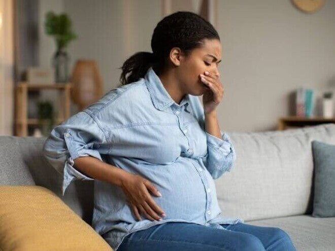 Enjoo na gravidez Gestante sentada no sofá com a mão na barriga e cobrindo a boca, demonstrando mal-estar, cenário comum para quem procura saber o que é bom para enjoo na gravidez.