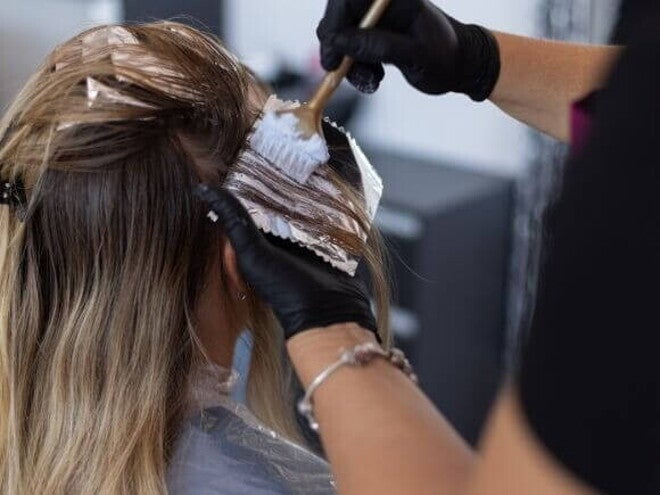 Mãos com luvas aplicando tinta no cabelo loiro de uma mulher, usando pincel e papel alumínio para mechas.