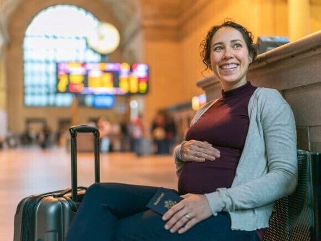 Foto de uma gestante sorrindo e relaxada, sentada em um banco de estação, com o passaporte na mão, indicando viagem internacional.