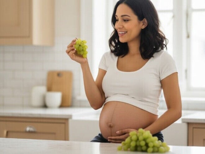 Mulher grávida com a mão na barriga segurando um cacho de uvas sentada em uma cozinha de tons claros