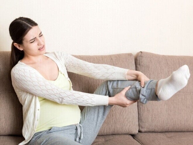 Gestante sentada no sofá, com expressão de dor, massageando a perna devido ao inchaço na gravidez comum nos membros inferiores.