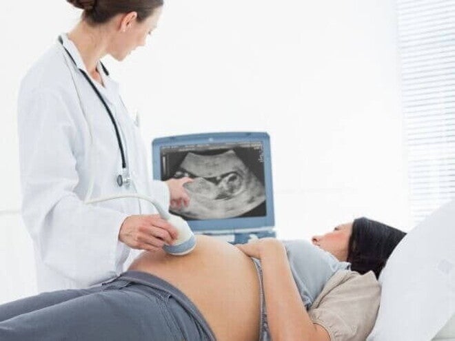 Médica realiza exame de ultrassonografia em mulher grávida para diagnóstico de líquido amniótico aumentado. A imagem mostra o monitor do aparelho exibindo o feto e a médica apontando para a tela.