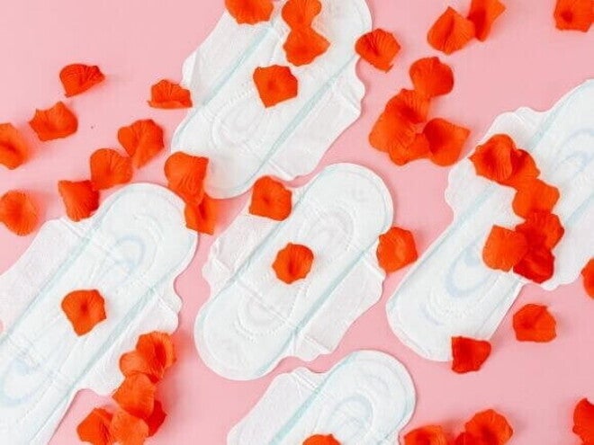 Quatro absorventes higiênicos dispostos sobre um fundo rosa, com pétalas de rosa vermelhas espalhadas.