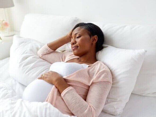 Mulher grávida deitada na cama, de lado, com os olhos fechados. A mão dela está na testa. A imagem mostra a posição para gestantes dormir, com a mulher buscando o conforto necessário para o sono.