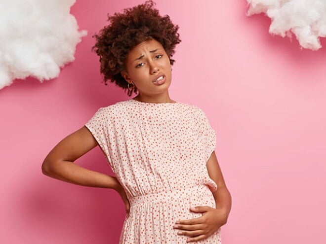 Mulher grávida em frente a uma parede rosa passando a mão na barriga e com expressão de desconforto. Mulher grávida em frente a uma parede rosa passando a mão na barriga e com expressão de desconforto.