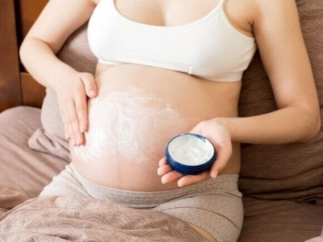Mulher grávida sentada na cama aplicando um creme hidratante branco na barriga para aliviar o prurido na gravidez. Ela segura um pote azul de creme em uma das mãos enquanto espalha o produto na pele esticada com a outra
