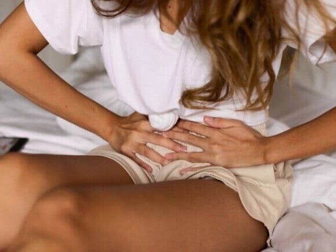 Mulher com blusa branca e shorts bege sentada com as mãos na parte inferior do abdômen, sentindo cólica Mulher com blusa branca e shorts bege sentada com as mãos na parte inferior do abdômen, sentindo cólica