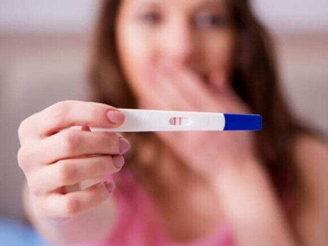 Mulher com expressão de surpresa, segurando um teste de gravidez que mostra o resultado positivo.