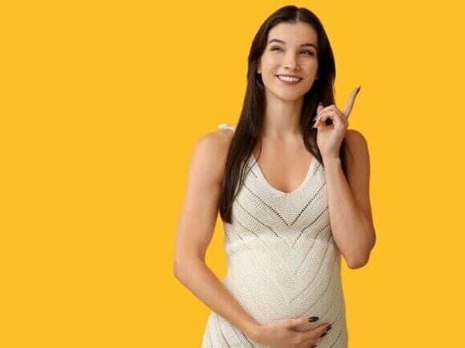 Mulher grávida sorridente, apontando para cima com o dedo. Ela usa um vestido branco e está de frente para um fundo amarelo. A imagem sugere a descoberta e o esclarecimento sobre os tipos de tampão mucoso durante a gravidez.