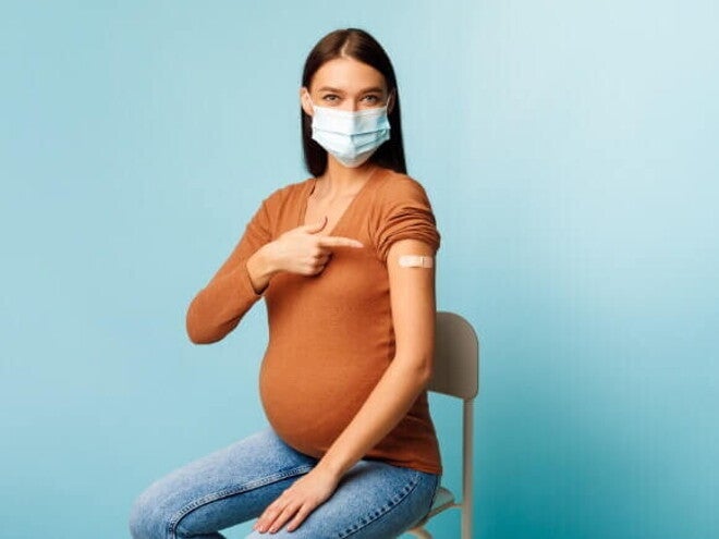 Mulher grávida com máscara apontando para o curativo no braço após receber uma das vacinas e gravidez recomendadas, simbolizando a proteção durante a gestação.