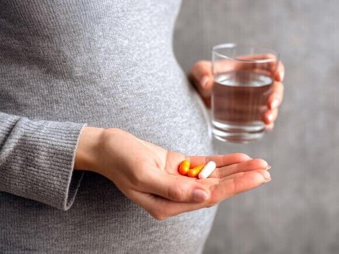 Mulher grávida, segurando na mão um copo de água e três cápsulas/pílulas, exemplificando a suplementação de vitaminas para gestante.