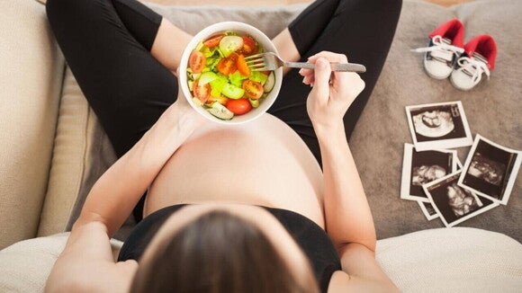 Mulher com 33 semanas de gravidez sentada, comendo uma salada, ao lado de fotos de ultrassom e sapatinhos de bebê.