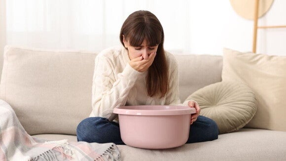 Mulher jovem sentada em um sofá segurando uma bacia rosa no colo e cobrindo a boca com a mão.