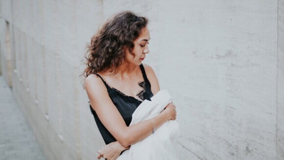 Mulher com cabelo cacheado vestindo blusa preta de renda, segurando um casaco branco, olhando discretamente para baixo.