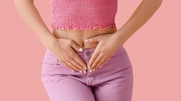 Mulher vestindo blusa rosa e calça roxa, com as mãos posicionadas sobre a parte inferior do abdômen, com desconforto por conta de cisto no ovário.