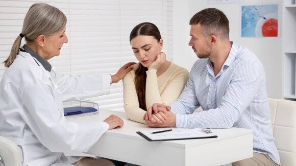 Casal sentado em uma consulta com uma médica de fertilidade, que toca o ombro da mulher de forma reconfortante enquanto discutem opções para aumentar a fertilidade e conceber.