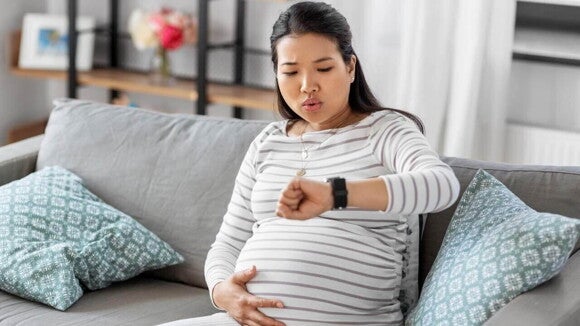 Mulher grávida, sentada em um sofá, olha para o relógio em seu pulso enquanto segura a barriga. A expressão facial dela indica que está monitorando a frequência e a intensidade das contrações de treinamento. Mulher grávida, sentada em um sofá, olha para o relógio em seu pulso enquanto segura a barriga. A expressão facial dela indica que está monitorando a frequência e a intensidade das contrações de treinamento.