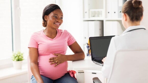 gestante sorrindo com a mão na barriga durante consulta médica, sentada em frente a profissional de saúde com notebook.