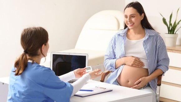 Gestante sorridente com as mãos sobre a barriga em um consultório médico com uma profissional de saúde segurando um glicosímetro à frente dela, simbolizando a atenção à diabete gestacional durante a gravidez.