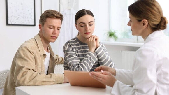 Casal jovem e caucasiano sentado em uma consulta, olhando para uma médica que lhes mostra informações em um tablet.