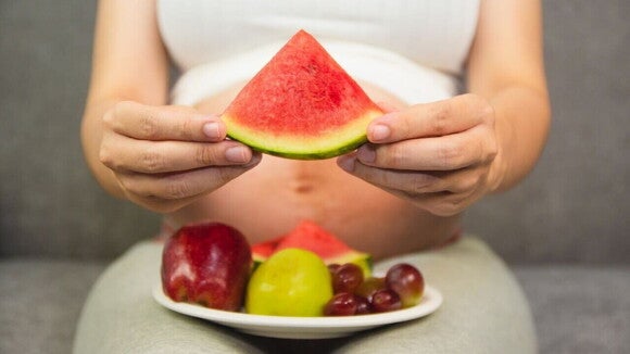 Mulher grávida com um prato de frutas no colo segurando uma fatia de melancia Mulher grávida com um prato de frutas no colo segurando uma fatia de melancia
