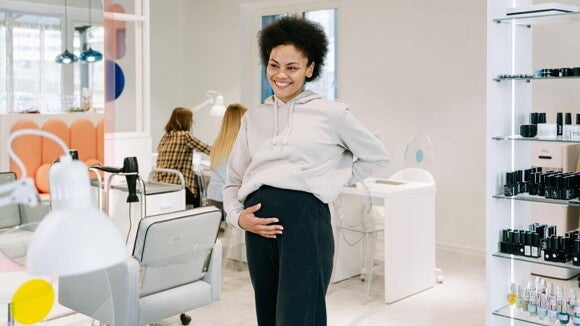 Mulher grávida sorrindo e segurando a barriga em um salão de beleza, cercada por prateleiras de esmaltes.