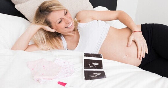 Mulher deitada em uma cama e sorrindo, com a mão na barriga, ao lado de um exame de ultrassom, um teste de gravidez e uma roupinha de bebê. Mulher deitada em uma cama e sorrindo, com a mão na barriga, ao lado de um exame de ultrassom, um teste de gravidez e uma roupinha de bebê.