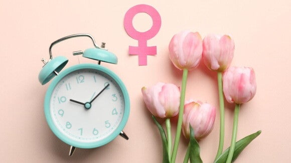 Composição com despertador azul, símbolo feminino rosa e tulipas sobre fundo claro, ilustrando o conceito de idade ideal para engravidar.