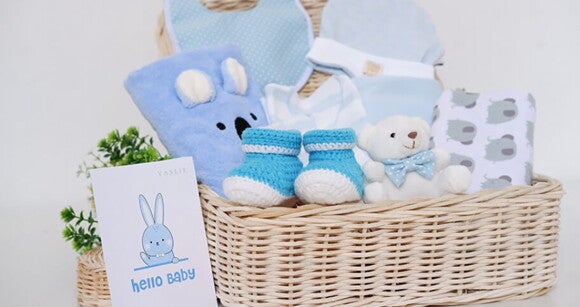 Imagem de uma cesta com vários itens de bebê, como cobertor, manta, sapatinho, babador, touca e um coelhinho de pelúcia Imagem de uma cesta com vários itens de bebê, como cobertor, manta, sapatinho, babador, touca e um coelhinho de pelúcia