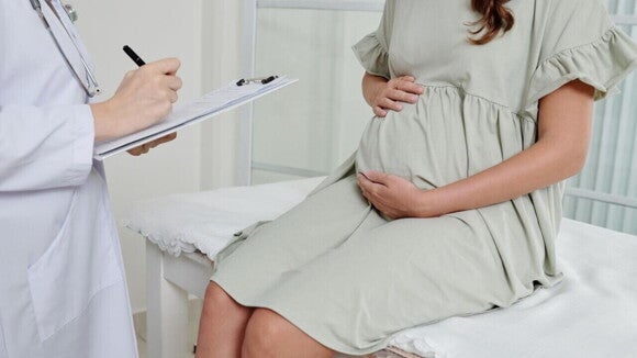 Médica com jaleco branco escreve em prontuário enquanto atende gestante sentada em consultório para avaliar casos de líquido amniótico aumentado. A paciente repousa as mãos sobre a barriga.