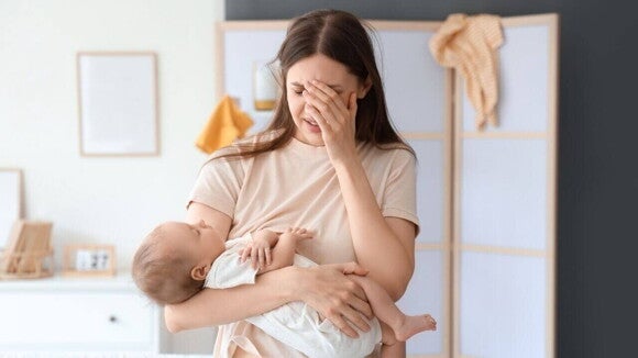 Uma mãe visivelmente exausta, com a mão no rosto, segurando seu bebê no colo em casa.