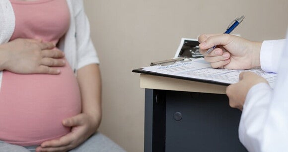 Médico escreve em uma prancheta enquanto uma mulher grávida está sentada à sua frente Médico escreve em uma prancheta enquanto uma mulher grávida está sentada à sua frente