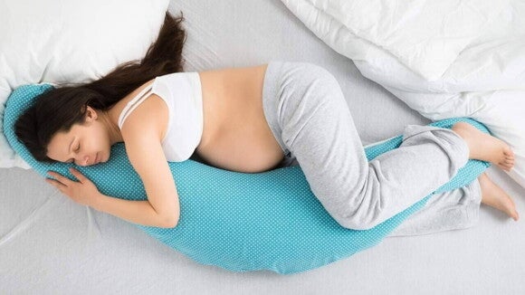 Mulher grávida, deitada de lado, dormindo confortavelmente em uma cama. Ela está com as mãos sobre a barriga, usando um travesseiro de corpo. A imagem exemplifica a posição para gestantes dormir.
