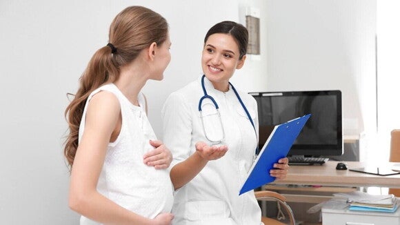 Uma gestante conversa e sorri com uma médica ou enfermeira em um consultório, representando o início dos cuidados e orientações do pré-natal.