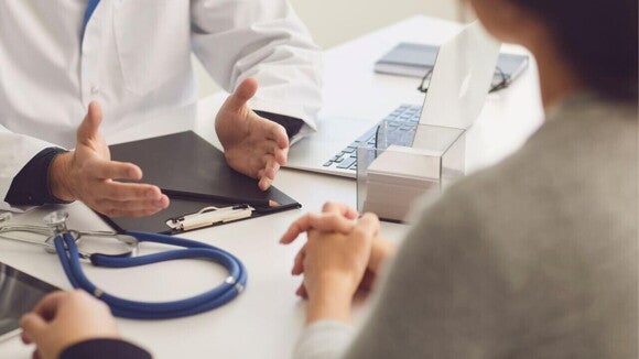 Homem com jaleco gesticula sentado em frente a uma mesa com estetoscópio, computador e outros equipamentos médicos, falando com um casal Homem com jaleco gesticula sentado em frente a uma mesa com estetoscópio, computador e outros equipamentos médicos, falando com um casal