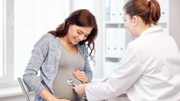 Uma médica usa um estetoscópio para ouvir o coração do feto na barriga de uma mulher grávida. A imagem ilustra a atenção médica durante a gravidez, abordando temas como sintomas de verme em gestante.