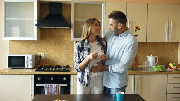Casal feliz na cozinha, mulher sorri enquanto mostra um teste de gravidez positivo.