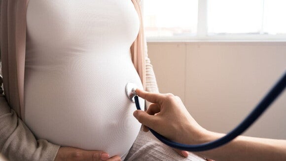 Profissional de saúde usa um estetoscópio para ouvir o coração do bebê na barriga de uma mulher grávida. A cena remete ao acompanhamento médico da gravidez, que pode envolver a identificação de diferentes tipos de tampão mucoso.