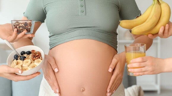 Close-up do abdômen de uma mulher grávida com várias mãos ao redor oferecendo alimentos saudáveis, representando a importância da nutrição e das vitaminas para gestante através da alimentação.