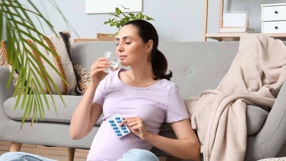 Mulher grávida, sentada no chão da sala, está prestes a tomar uma pílula de uma cartela de medicamentos enquanto segura um copo de água. A imagem representa o momento de ingerir as vitaminas pré-natais.
